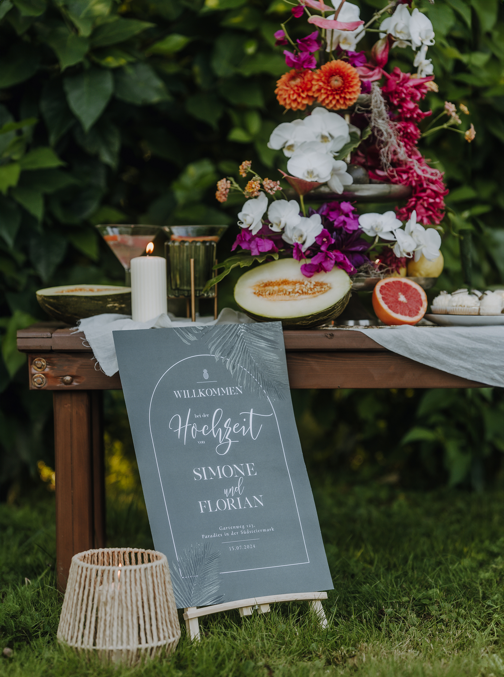 Dekorierter Tisch bei einer Gartenhochzeit mit Früchten, Blumen und einem Willkommensschild.